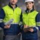 Two site engineers in yellow and navy hi-vis jackets with white hard hats